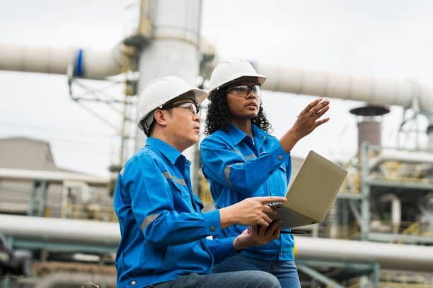 Engineers in hard hats and blue uniforms inspect industrial facility using a laptop.