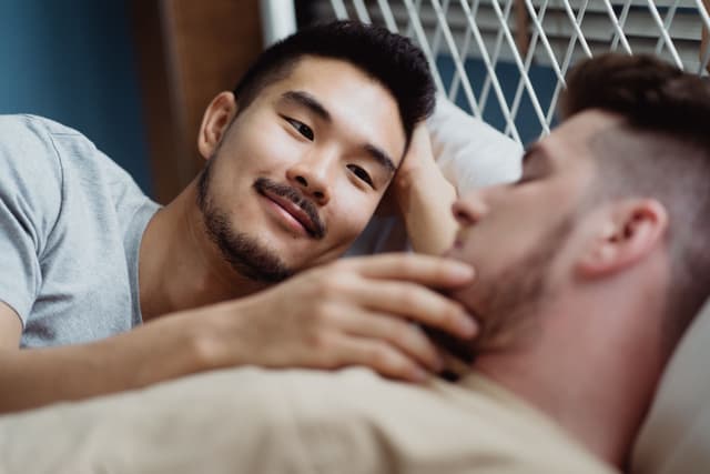 Asian man smiles at viewer while gently touching a bearded man's chin.