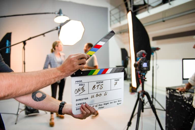 Hands hold a clapperboard on a film set with lights and people in the blurred background.