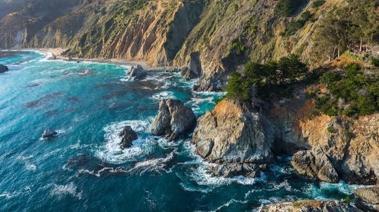 Aerial view of a rugged coastline with turquoise ocean, towering cliffs, and prominent sea stacks.