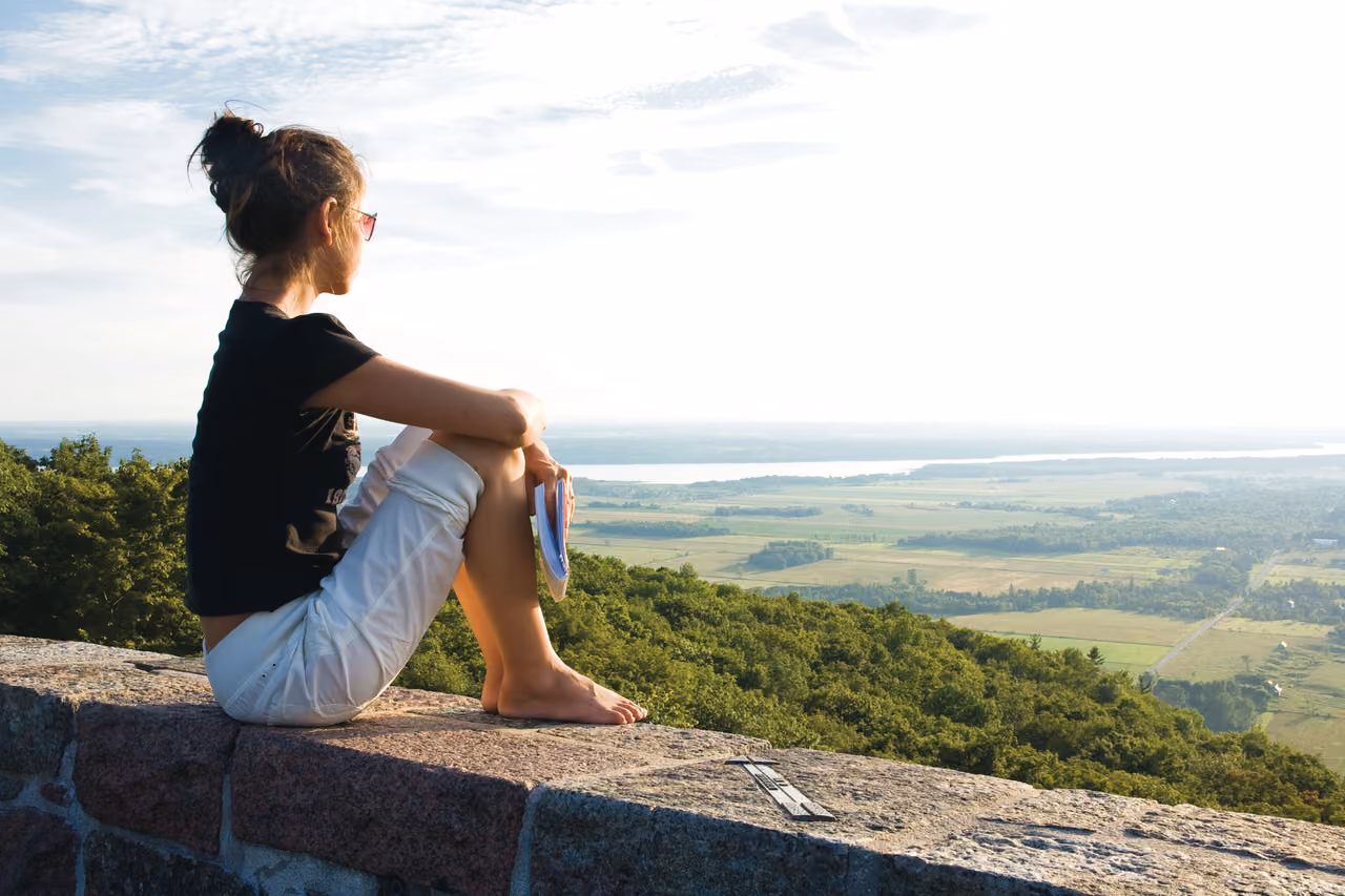 Person sitting on a stone wall, contemplating a vast landscape with a river and fields.