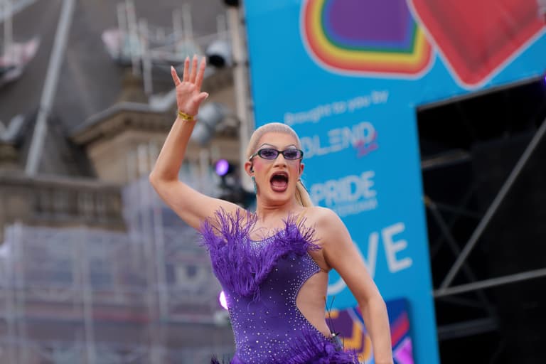 Blonde woman in purple sparkly dress with feathers singing on stage at Pride event.